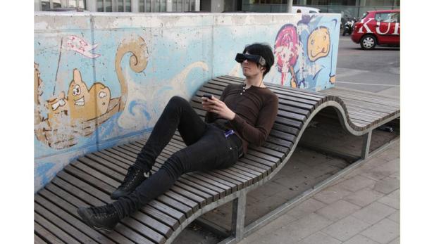 Ein Mann mit VR-Brille liegt auf einer geschwungenen Holzbank und benutzt sein Smartphone.