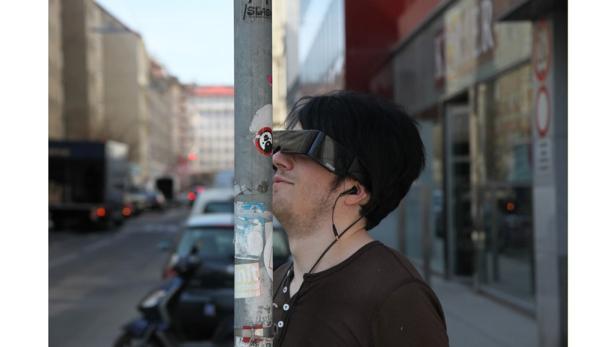 Ein Mann mit schwarzen Haaren und futuristischer Brille steht an einem Laternenpfahl.