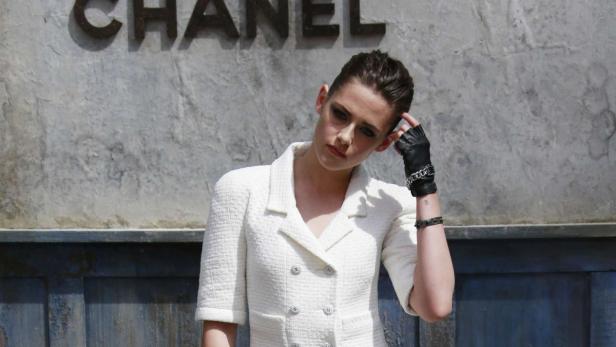 Kristen Stewart vor einem Chanel-Logo mit weißem Blazer und schwarzen Handschuhen.