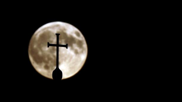 Ein Kreuz ragt vor einem hellen Vollmond in den Nachthimmel.