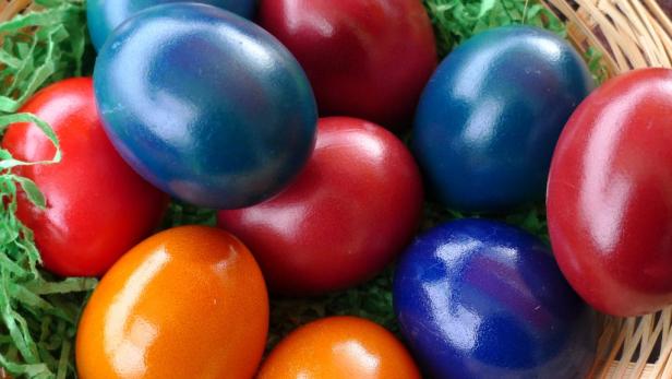 Einige rot, blau und orange gefärbte Ostereier liegen in einem Korb mit grünem Papiergras.