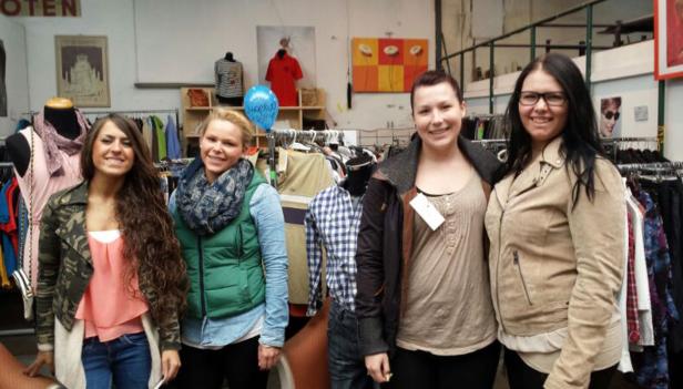 Vier Frauen posieren in einem Secondhand-Laden vor Kleiderständern.