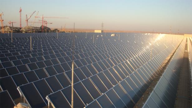 Ein Solarkraftwerk erstreckt sich bis zum Horizont, im Hintergrund Baustellenkräne.
