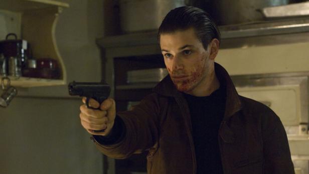 Ein Mann mit Blut im Gesicht zielt mit einer Pistole.