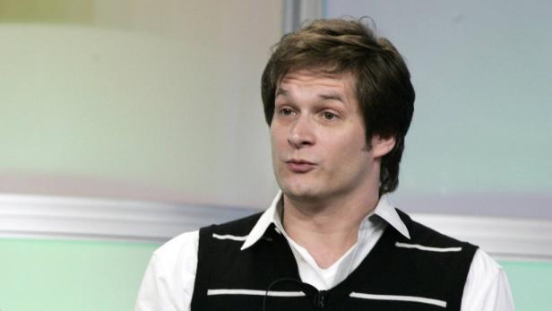 Ein Mann mit braunen Haaren und einem schwarzen Pullunder mit weißen Streifen.