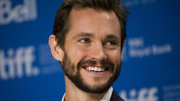 Porträt des lächelnden Schauspielers Hugh Dancy vor einem blauen Hintergrund.