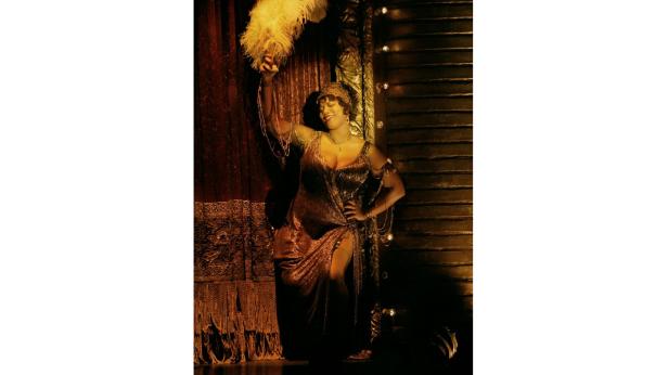 Eine Frau in einem goldenen Kleid posiert mit einem Federfächer auf einer Bühne.