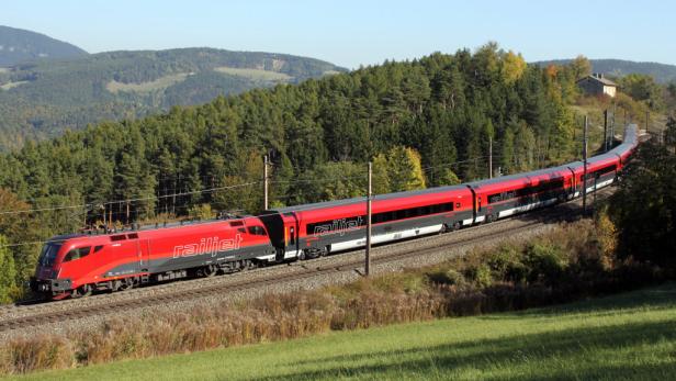 Ein roter Railjet-Hochgeschwindigkeitszug fährt durch eine grüne Landschaft.