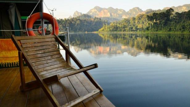 Ein leerer Holzliegestuhl steht am Ufer eines ruhigen Sees mit Bergen im Hintergrund.