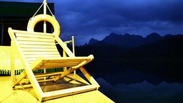 Ein Liegestuhl und ein Rettungsring stehen auf einem Steg mit Blick auf einen dunklen See und Berge.