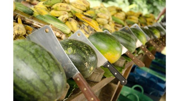 Auf einem Marktstand liegen Wassermelonen und Bananen mit Hackmessern zum Verkauf bereit.