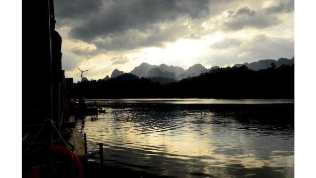 Dunkle Silhouette einer bergigen Landschaft am Ufer eines Sees unter bewölktem Himmel.