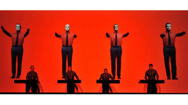 Die Band Kraftwerk tritt vor einem roten Hintergrund auf, auf dem vier Schaufensterpuppen stehen.