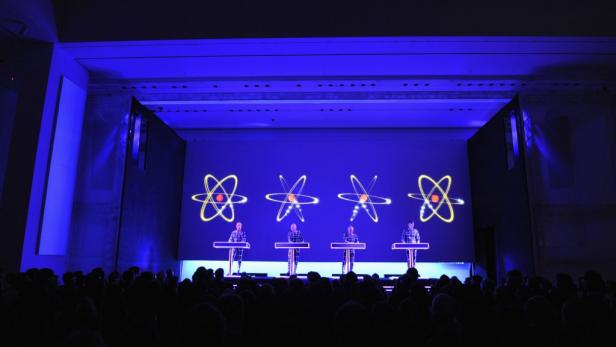 Die Band Kraftwerk tritt vor einem Publikum mit Atomgrafiken im Hintergrund auf.
