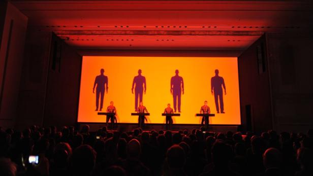 Die Band Kraftwerk tritt vor einem großen Publikum auf einer Bühne mit orangefarbenem Hintergrund auf.