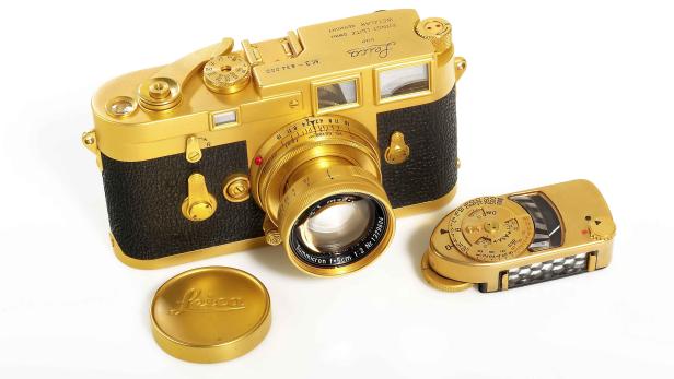 Eine goldene Leica M3 Kamera mit Belichtungsmesser und Objektivdeckel auf einem weißen Hintergrund.