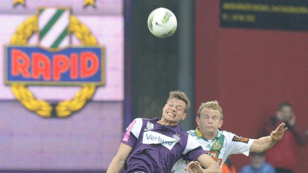 Zwei Fußballspieler kämpfen um einen Ball in der Luft vor dem Rapid-Logo.