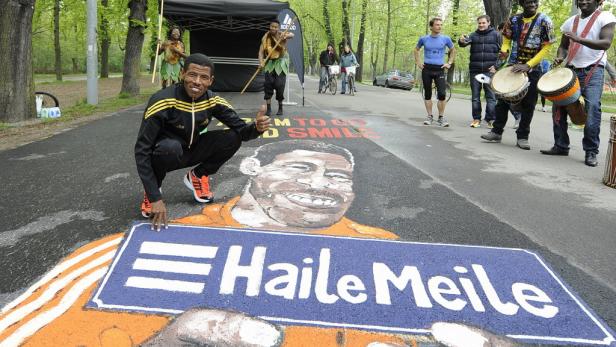 Haile Gebrselassie posiert vor einem Straßenkunstwerk, das ihn und den Schriftzug „Haile Meile“ zeigt.
