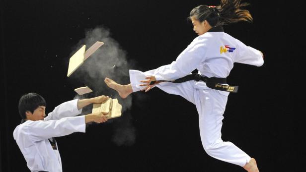 Eine Frau im Taekwondo-Anzug zerschlägt mit einem Tritt ein Holzbrett, das von einem Mann gehalten wird.