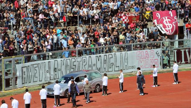 Ein Leichenwagen auf einem Fußballfeld, umgeben von Trauernden und Transparenten.
