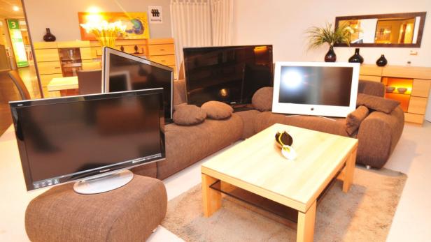 Mehrere Fernseher stehen auf einem braunen Sofa und einem Hocker in einem Wohnzimmer.