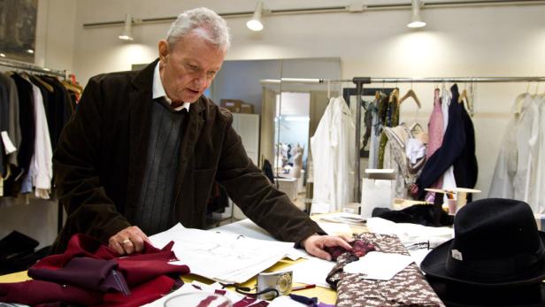 Ein älterer Mann beugt sich über einen Tisch mit Stoffen und Skizzen in einem Atelier.