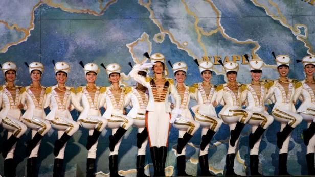 Eine Gruppe von Tänzerinnen in weißen Uniformen und schwarzen Stiefeln tritt auf einer Bühne auf.