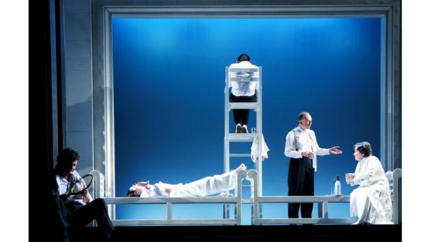 Eine Theaterszene mit mehreren Schauspielern auf einer Bühne vor blauem Hintergrund.