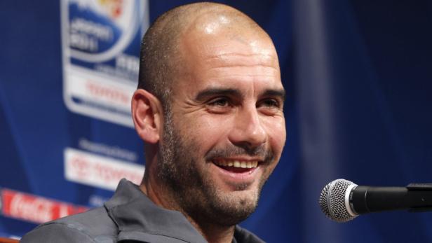 Pep Guardiola lächelt vor einem Mikrofon bei einer Pressekonferenz.