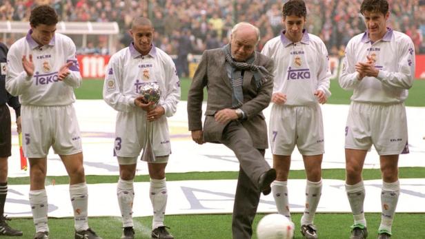 Ein älterer Mann im Anzug schießt einen Fußball vor einer Gruppe von Real Madrid-Spielern.