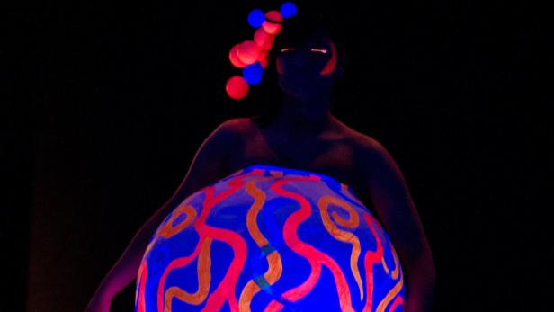 Eine Person im Dunkeln mit leuchtenden Farben und Kugeln im Haar präsentiert ein blaues, leuchtendes Objekt.