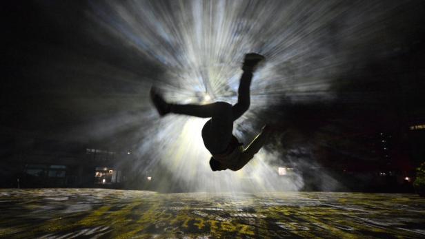 Eine Person macht einen Salto vor einer hellen Lichtinstallation im Dunkeln.