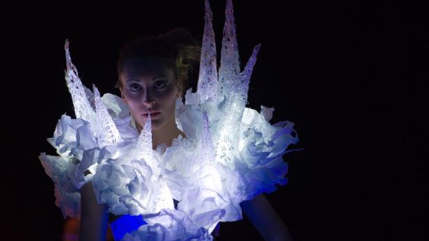 Eine Frau trägt ein leuchtendes, futuristisches Kleid mit spitzen Verzierungen.