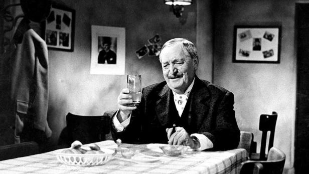 Ein älterer Mann mit Schnurrbart sitzt an einem Tisch und hält ein Glas in der Hand.