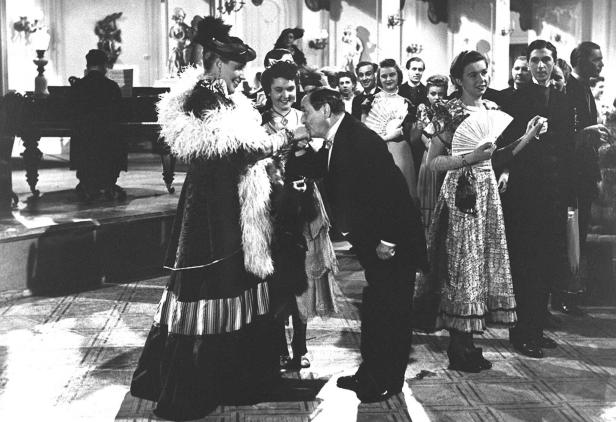 Ein Mann küsst die Hand einer elegant gekleideten Frau auf einem Ball.