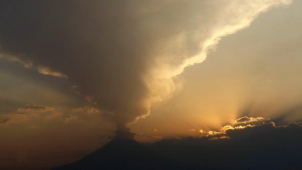 Ein Vulkan ragt unter einem dramatischen Himmel hervor, durch den Sonnenstrahlen dringen.
