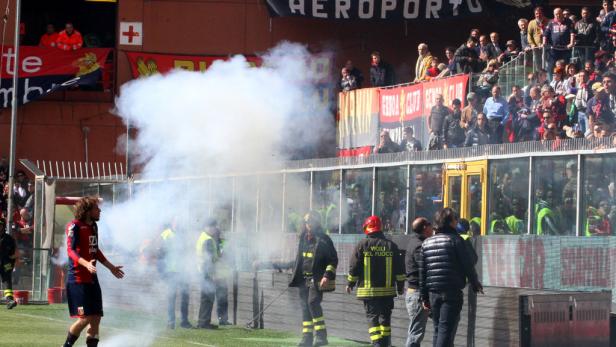 Ein Fußballspieler gestikuliert, während Feuerwehrleute Rauch auf dem Spielfeld beseitigen.