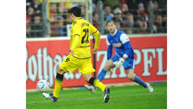 Ein Fußballspieler in gelber Uniform schießt den Ball, während ein Torwart in Blau zusieht.