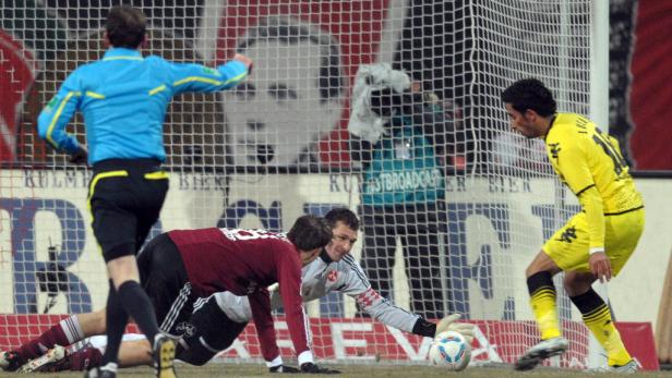 Ein Fußballtorwart versucht, einen Schuss abzuwehren, während ein Schiedsrichter zusieht.