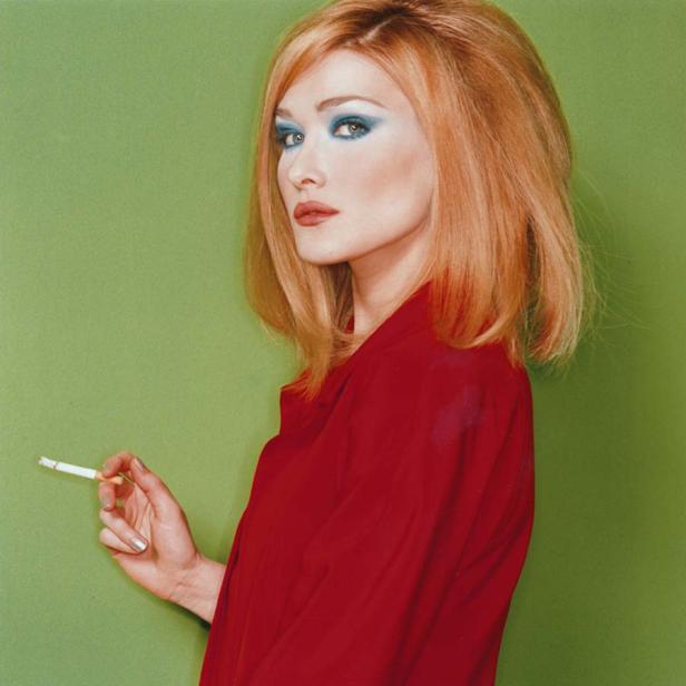 Eine Frau mit rotem Haar und blauem Lidschatten raucht eine Zigarette vor einem grünen Hintergrund.