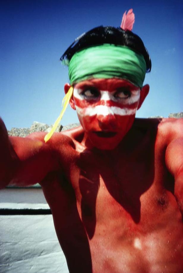 Ein Mann mit roter Farbe im Gesicht und einem grünen Stirnband vor blauem Himmel.