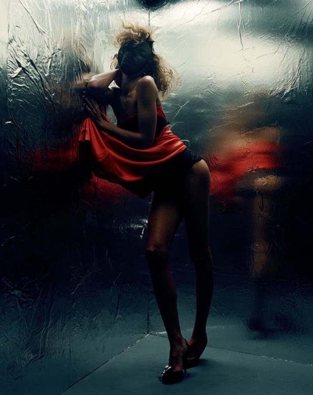 Eine Frau mit Maske und rotem Kleid posiert vor einer silbernen Wand.