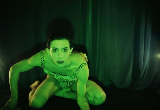 Ein Mann in einem Leopardenmuster-Outfit kniet vor einem grünen Vorhang.