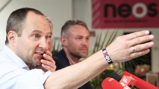 Ein Mann mit einem „neos“-Armband gestikuliert vor Mikrofonen.
