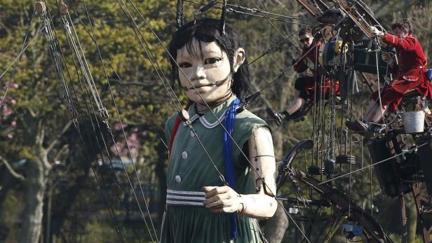 Eine riesige Marionette in Form eines Mädchens wird von mehreren Personen mit Seilzügen bewegt.