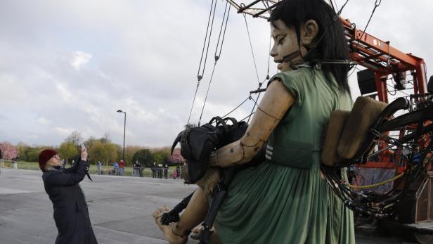 Eine Frau fotografiert eine riesige, sitzende Marionette mit dunklen Haaren und grünem Kleid.