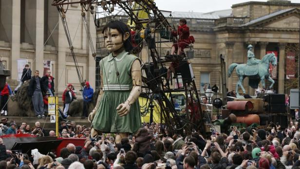 Eine riesige Marionette in Form eines Mädchens wird von einer Menschenmenge bestaunt.