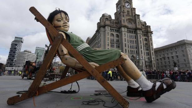 Eine riesige Marionette in einem grünen Kleid sitzt in einem Liegestuhl vor dem Royal Liver Building in Liverpool.
