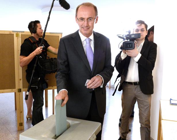 Ein Mann im Anzug wirft einen Stimmzettel in eine Wahlurne, gefilmt von Kameraleuten.