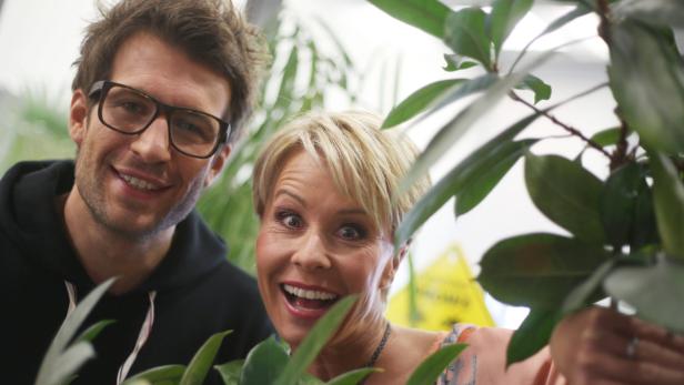 Ein Mann mit Brille und eine Frau mit kurzem blonden Haar schauen lächelnd zwischen grünen Pflanzen hervor.
